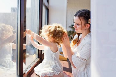 Anne ve kızı pencere eşiğinde pencereden dışarı bakıyor. Büyük bir pencerenin arkasında mutlu bir aile var. Evde kal. Ev Karantinası