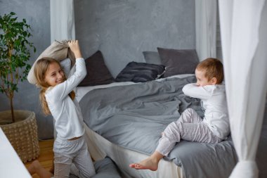Yaramaz çocuklar pijamalı bir kız ve küçük bir çocuk gri bir yatak odasındaki yatakta yastık savaşı yaptılar. Bu oyunu seviyorlar. Ev eğlencelidir..