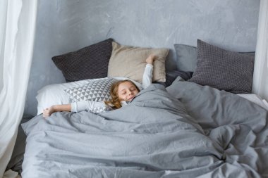 Çocuk çalar saatle gri tonlarda bir yatakta uyuyor. Uyanış.