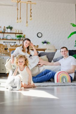 Neşeli küçük bir kız Sunny oturma odasında doldurulmuş fil oyuncağıyla oynuyor. Ebeveynler arka planda bulanık oturuyorlar. Evimiz çok sıcak..