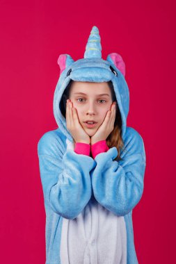 Çekici bir kız tek boynuzlu at kostümü içinde şakacı bir şekilde poz veriyor. Kigurumi 'de elleri pembe bir arka planda duran şaşırmış bir kızın stüdyo fotoğrafı..