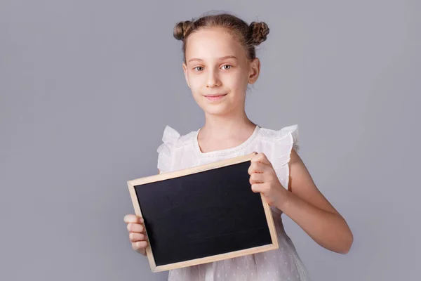 少女は灰色の背景に空のボードを保持しています 子は空のボードを表示します 教育の概念 — ストック写真