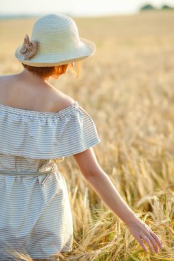 Kafasında hasır şapka olan genç bir kız buğday tarlasında dans ediyor. Elini kulakların üzerinden geçiriyor. Sırtı kameraya dönük duruyor..