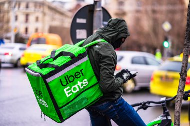 Uber Eats logosuna sahip bisikletli genç adam, Romanya 'nın başkenti Bükreş' te yağmurlu bir günde yemek dağıtıyor.
