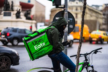 Uber Eats logosuna sahip bisikletli genç adam, Romanya 'nın başkenti Bükreş' te yağmurlu bir günde yemek dağıtıyor.