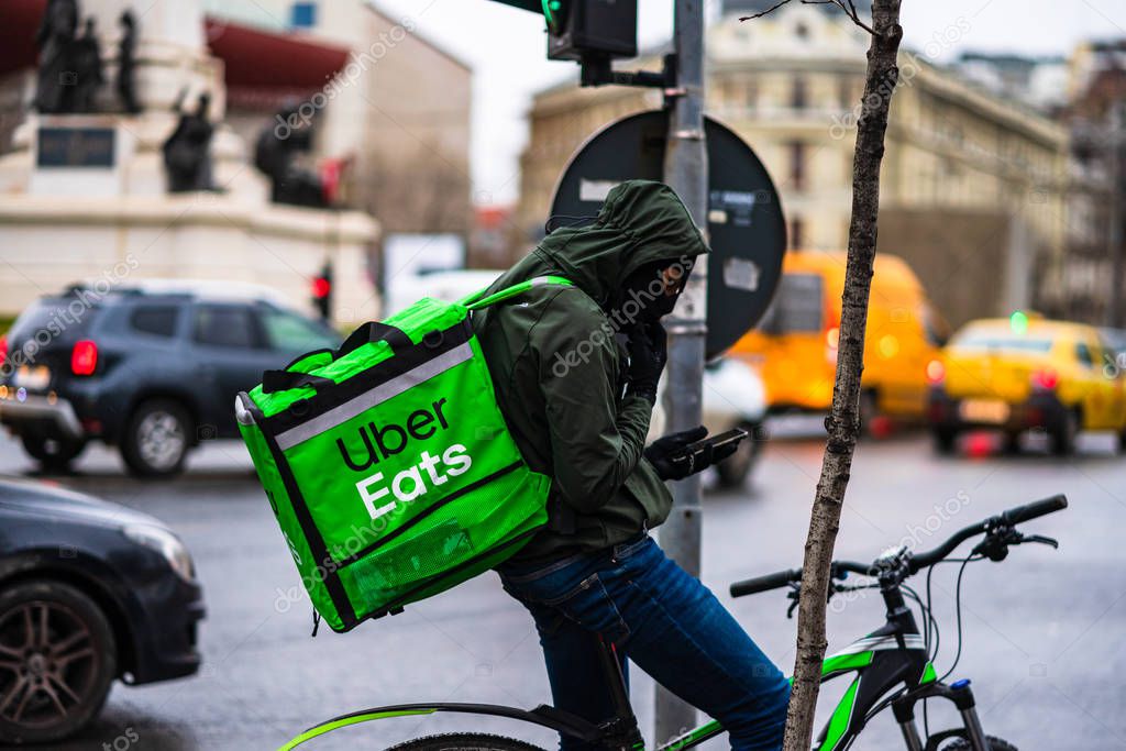 Young man on a bike with Uber Eats logo delivering food during a rainy day in Bucharest, Romania, 2020