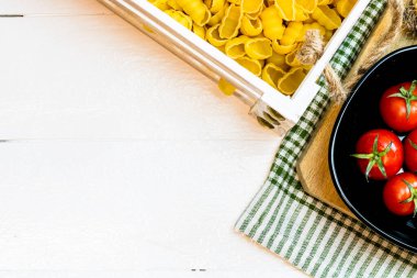 Tahta bir sandıkta makarna ve kırsal beyaz tahta bir masada siyah bir kasede taze kiraz domatesleri. Malzemeler ve gıda konsepti