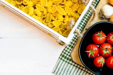 Soğan, sarımsak, ahşap bir sandıkta makarna ve kırsal beyaz ahşap bir masada taze olgun domates manzarası. Malzemeler ve gıda konsepti