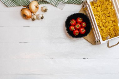 Soğan, sarımsak, ahşap bir sandıkta makarna ve kırsal beyaz ahşap bir masada taze olgun domates manzarası. Malzemeler ve gıda konsepti