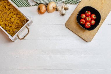 Soğan, sarımsak, ahşap bir sandıkta makarna ve kırsal beyaz ahşap bir masada taze olgun domates manzarası. Malzemeler ve gıda konsepti