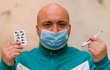 Tıbbi maskeli hasta genç adam portreyi kapat. Adamın elinde haplar ve bulanık arka planda termometre var. Covid-19 salgın kirliliği kavramı