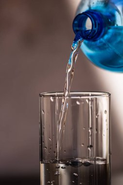 Mavi plastik şişeden bulanık arka plandaki bir bardağa su dökülüyor. Seçici odak, sığ DOF ve kopyalama alanı