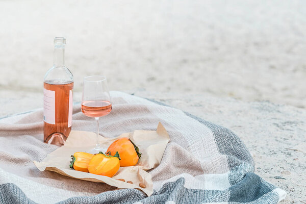 Picnic on the beach with persimmons, almond and bottle of rose wine on beige blanket.