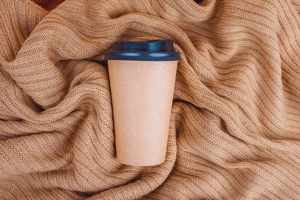 Coffee paper cup on beige knitted blanket background, top view flat lay. Take away coffee cup.