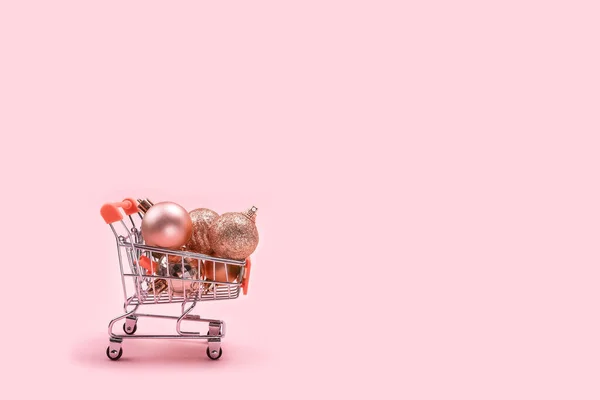 Christmas tree decorations and balls in miniature shopping cart with on pastel pink background with copy space. — Stock Photo, Image