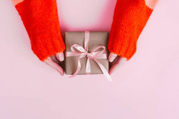 Frauenhände in warmem Strickpullover mit Maniküre halten Geschenkbox mit Bastelpapier und rosa Schleife auf rosa Hintergrund umwickelt. — Stockfoto