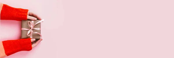 Frauenhände mit Maniküre halten Geschenkverpackung in Bastelpapier mit rosa Schleife auf blassrosa Hintergrund gewickelt. Festliches Konzept. — Stockfoto