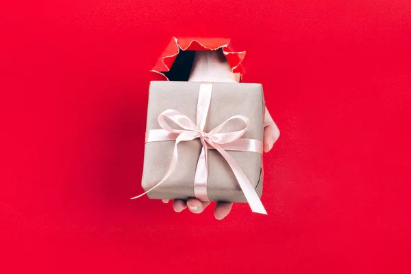 Female hand holding gift box wrapped in craft paper with pink ribbon through a hole in red background.