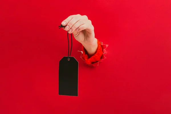Etiqueta negra en blanco en la mano femenina a través de un agujero en el fondo rojo. Etiqueta de precio, etiqueta regalo, etiqueta de dirección . — Foto de Stock