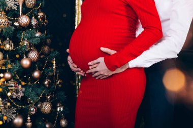 Anne ve babanın elleri, altın oyuncaklarla süslenmiş Noel ağacının yanında hamile karnına dokunuyor. Kırpılmış.