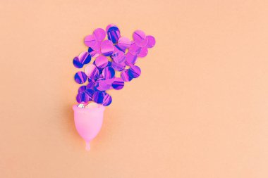 Pastel turuncu arka planda adet dönemini simgeleyen holografik pullu adet bardağı..