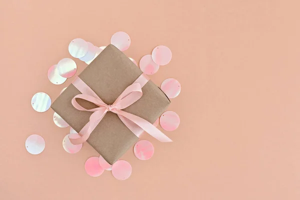 Caja de regalo envuelta en papel artesanal con cinta rosa sobre fondo naranja pálido con purpurina holográfica . — Foto de Stock