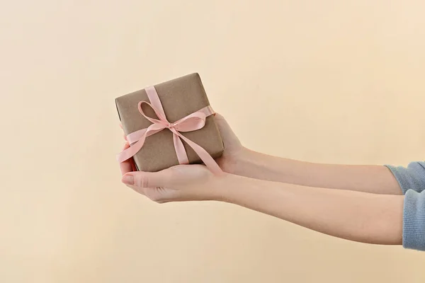 Weibliche Hand hält Geschenkschachtel in Bastelpapier eingewickelt mit rosa Schleife durch auf hellbeigem Hintergrund. Stockbild
