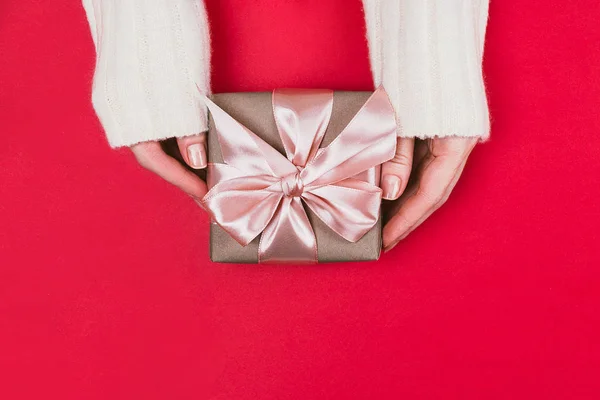 Las manos de la mujer sostienen la caja de regalo envuelta con papel artesanal y cinta rosa sobre fondo rojo . — Foto de Stock