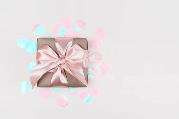 Pudełko zapakowane w papier rzemieślniczy z idealną różową wstążką na bladoróżowym rustykalnym tle z holograficznym konfetti. — Zdjęcie stockowe