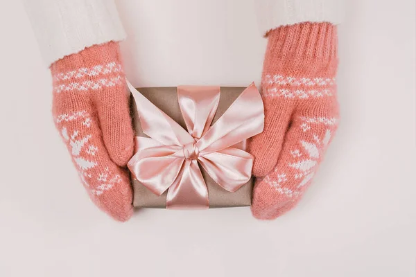 Frauenhände in warmen Strickhandschuhen halten Geschenkverpackung mit Bastelpapier und rosa Schleife auf weißem Hintergrund. — Stockfoto