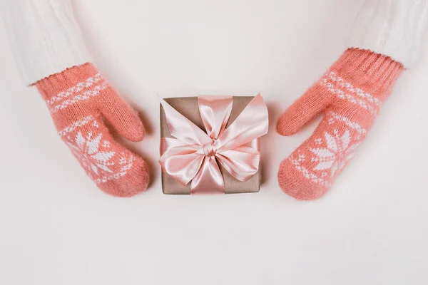 Frauenhände in warmen Strickhandschuhen halten Geschenkverpackung mit Bastelpapier und rosa Schleife auf weißem Hintergrund. — Stockfoto