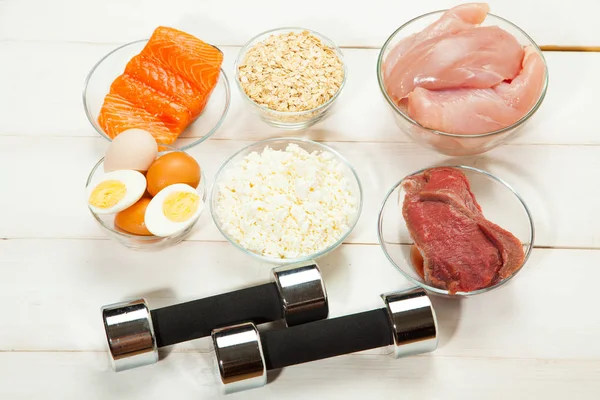 Dieta proteica, pesce, formaggio, uova, carne, pollo e manubri su uno sfondo di legno bianco — Foto Stock