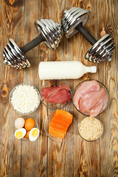 Protein, fish, cheese, eggs, meat, chicken and dumbbells on a wooden background