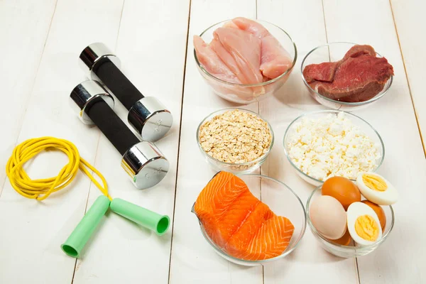 Dieta proteica, pesce, formaggio, uova, carne, pollo e manubri su uno sfondo di legno bianco — Foto Stock