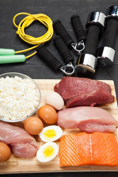 Proteínas y mancuernas. Pescado, queso, huevos, carne, corazón de pollo sobre un fondo de madera negro — Foto de Stock