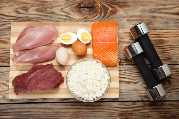 Dieta proteica, pescado, queso, huevos, carne, pollo y pesas sobre un fondo de madera — Foto de Stock