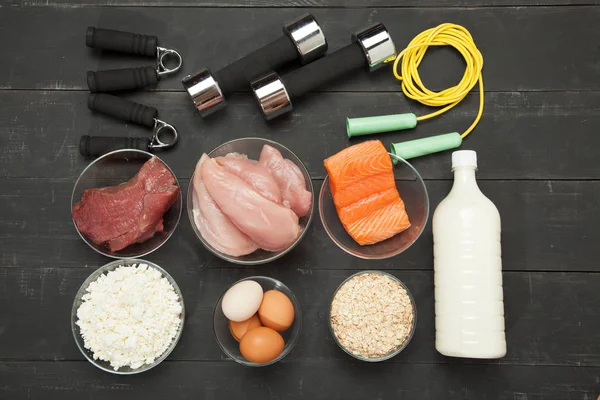 Proteínas y mancuernas. Pescado, queso, huevos, carne, corazón de pollo sobre un fondo de madera negro — Foto de Stock