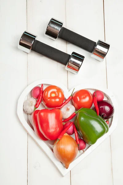 Sport e dieta. Uno stile di vita sano. Verdure, manubri. Peperoni, pomodori, aglio, ravanello di cipolla in un cuore su sfondo bianco — Foto Stock