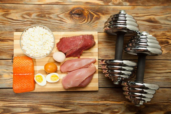 Dieta proteica, pescado, queso, huevos, carne, pollo y pesas sobre un fondo de madera — Foto de Stock