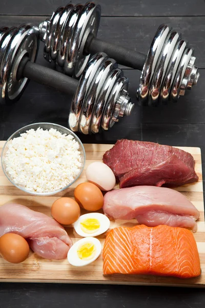 Proteínas y mancuernas. Pescado, queso, huevos, carne, corazón de pollo sobre un fondo de madera negro — Foto de Stock