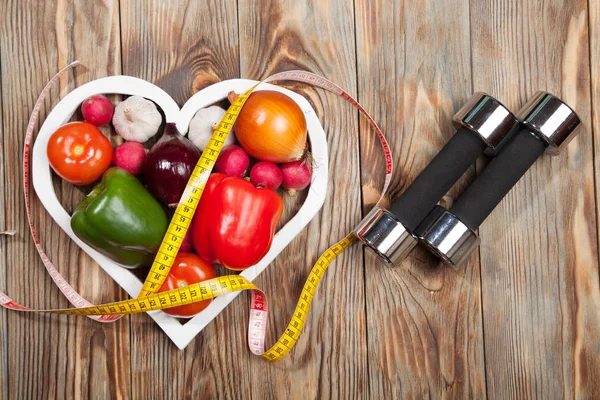 Sport en dieet. Groenten, halters centimeter. Paprika's, tomaten, knoflook, uien, radijsjes in het hart aan rustieke achtergrond Rechtenvrije Stockfoto's