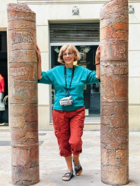 Beautiful mature happy tourist woman with old video camera on her neck travelling in Palma de Majorca stays between two ancient columns clipart