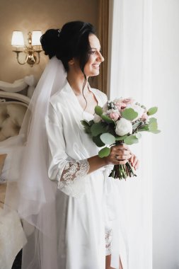 Genç ve güzel gelinin buketli portresi. Düğün günü toplantıları. Güzel bir kız, damadı beklerken bir otel odasında düğün töreni için giyinir ve toplanır. Gelinin sabahı