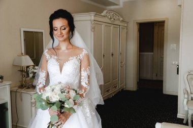 Genç ve güzel gelinin buketli portresi. Düğün günü toplantıları. Güzel bir kız, damadı beklerken bir otel odasında düğün töreni için giyinir ve toplanır. Gelinin sabahı