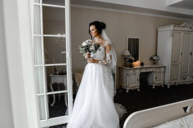 Genç ve güzel gelinin buketli portresi. Düğün günü toplantıları. Güzel bir kız, damadı beklerken bir otel odasında düğün töreni için giyinir ve toplanır. Gelinin sabahı