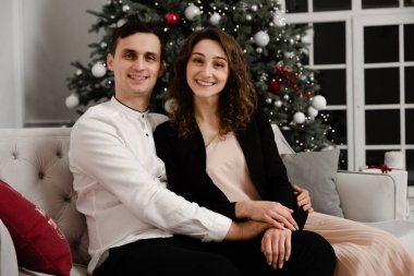 Genç çift kucaklaşıp kanepede oturuyor. Noel ağacının altında hediye kutuları. Güzel, modern bir dairede yeni yıl dekorasyonu. Tatil kavramı