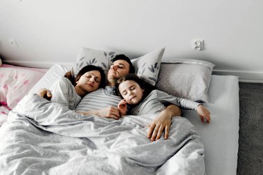 Anne babası yatakta, baba karısını ve kızını nazikçe öpüyor, güçlü aile kucaklaşmaları, aile hafta sonu yatağı, uyuyan aile, kız çocuğu yatakta genç anne babasıyla uyuyor.