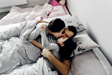 Anne babası yatakta, baba karısını ve kızını nazikçe öpüyor, güçlü aile kucaklaşmaları, aile hafta sonu yatağı, uyuyan aile, kız çocuğu yatakta genç anne babasıyla uyuyor.