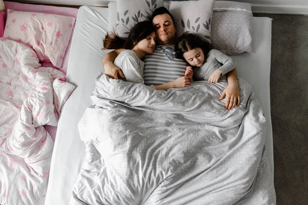 daughter with parents in bed,dad gently kisses his wife and daughter,strong family hugs,family weekend in bed,sleeping family,daughter sleeps with young parents in bed