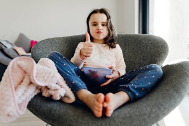 Bir kase yemekle bir sandalyede oturan güzel bir kız, tatlı bir bebek bir tabaktan yemek yerken bir kanepede oturan, pijamalı genç bir kız yemek yiyor.